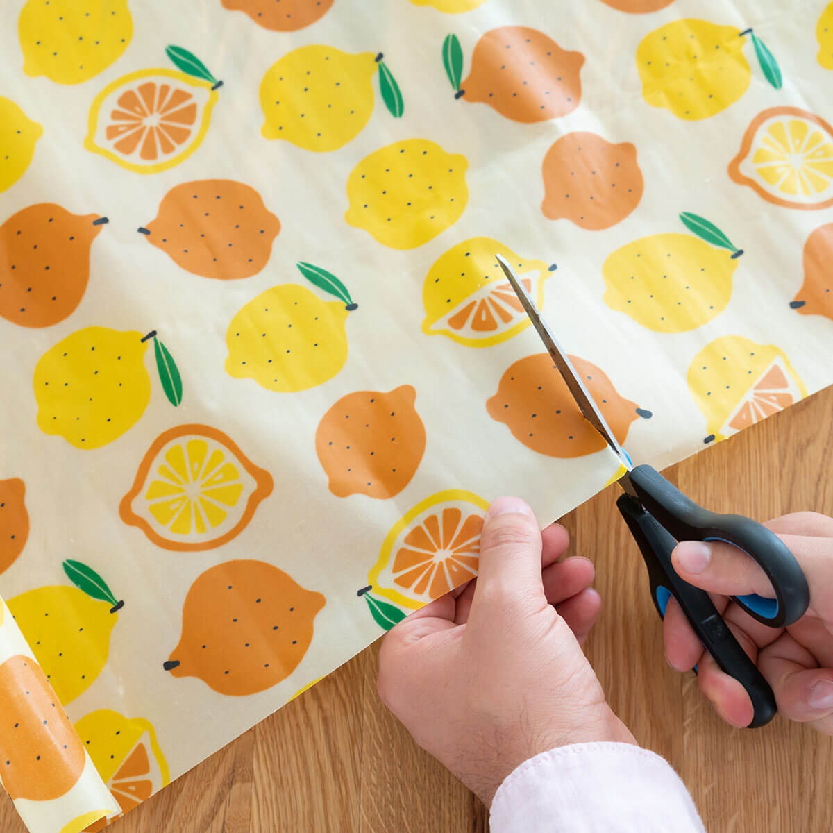 Hände schneiden ein bunt bedrucktes Bienenwachstuch mit Zitronen- und Orangenmotiv von Io Nova auf einem Holztisch mit einer Schere zurecht.