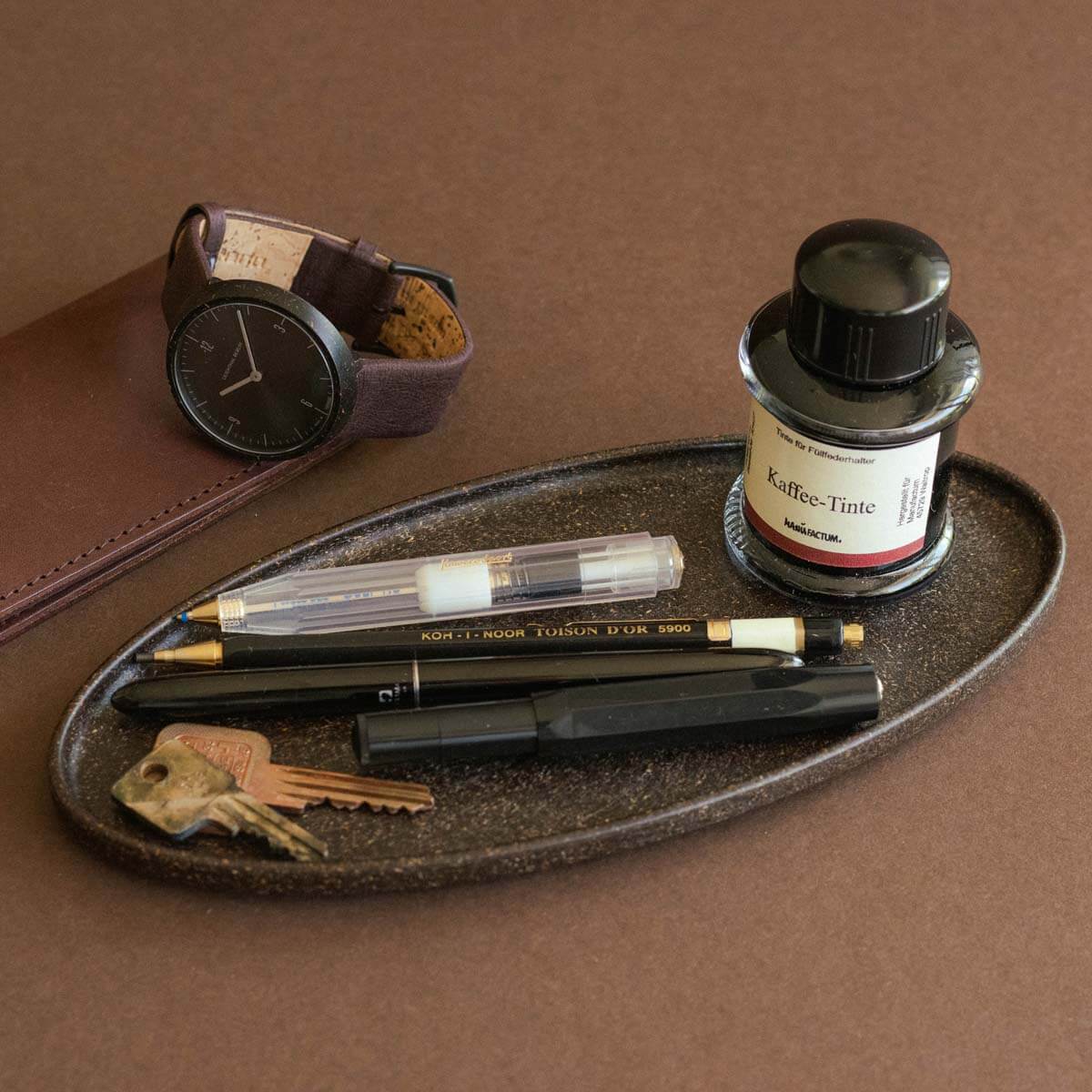 Braunes Tablett aus recyceltem Kaffeesatz mit Schreibutensilien, Schlüsseln und Kaffee-Tinte, stilvoll arrangiert auf braunem Untergrund.