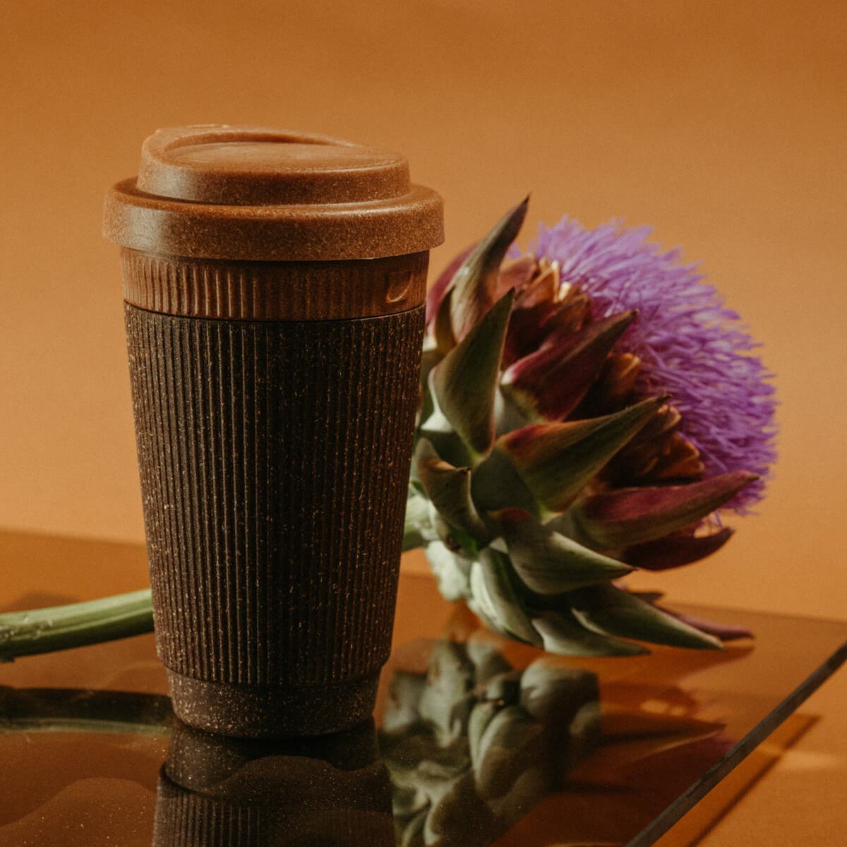 Wiederverwendbarer Kaffeebecher von Kaffeeform mit Deckel in Muskatfarbe, stehend auf einer reflektierenden Fläche neben einer lila Blüte.