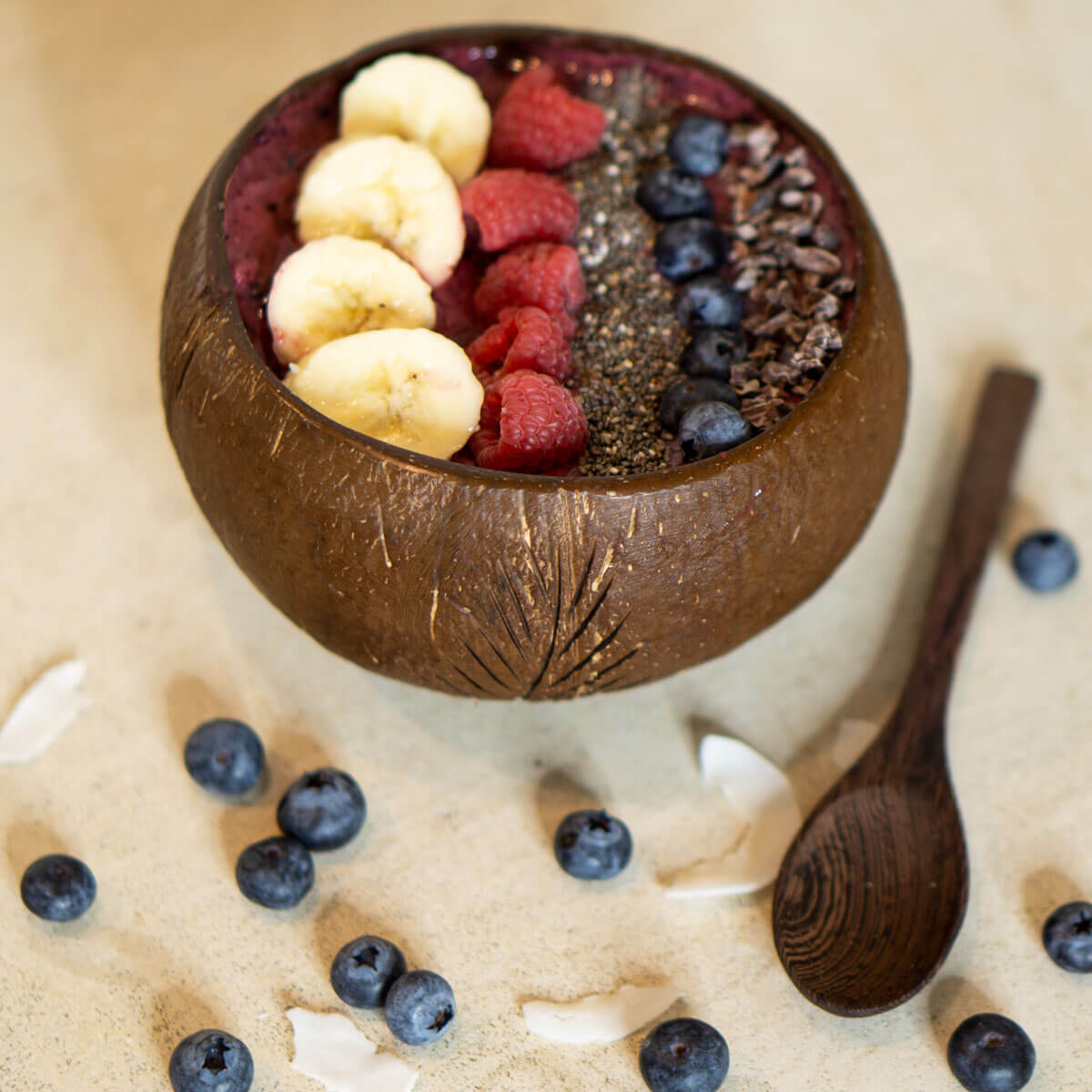 Kokosnussschale mit Smoothie-Bowl, belegt mit Banane, Himbeeren, Chiasamen, Blaubeeren und Kakaonibs auf heller Unterlage mit Holzlöffel.