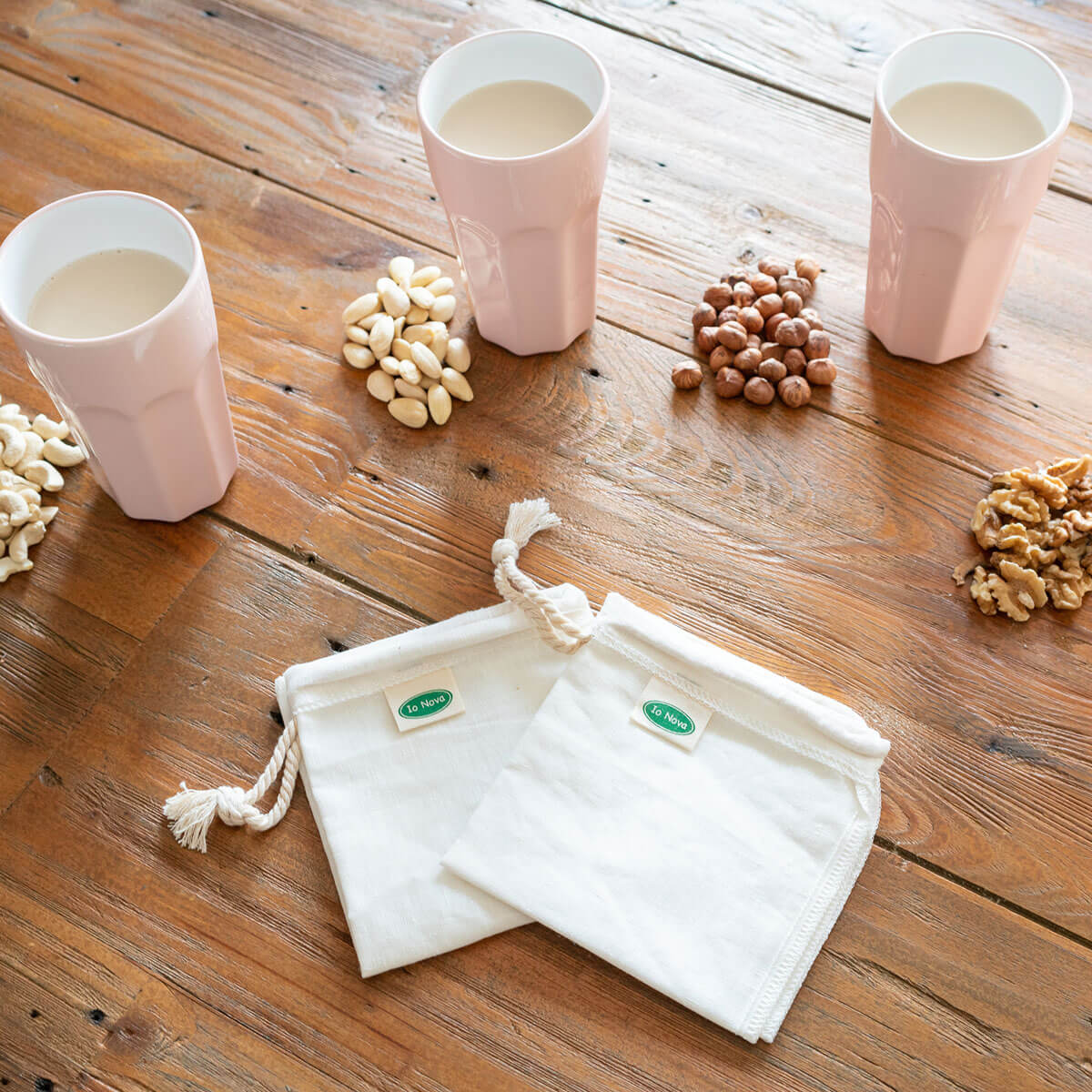 Zwei wiederverwendbare Nussmilchbeutel aus Hanffaser von Io Nova auf einem Holztisch, daneben rosa Gläser mit Pflanzenmilch und verschiedene Nuss-Sorten wie Mandeln, Haselnüsse, Cashews und Walnüsse.