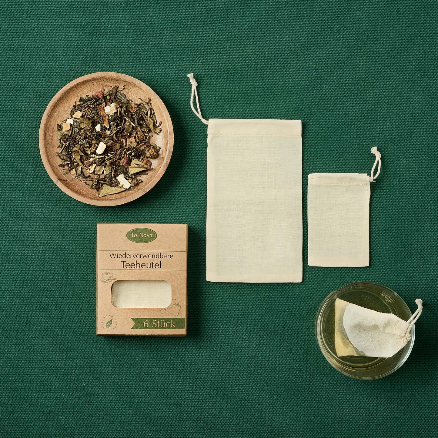 Wiederverwendbare Teebeutel aus Baumwolle von Io Nova auf grünem Stoffhintergrund – mit Verpackung, zwei verschiedenen Beutelgrößen, einer Schale mit losem Tee und einer Tasse mit eingelegtem Teebeutel.
