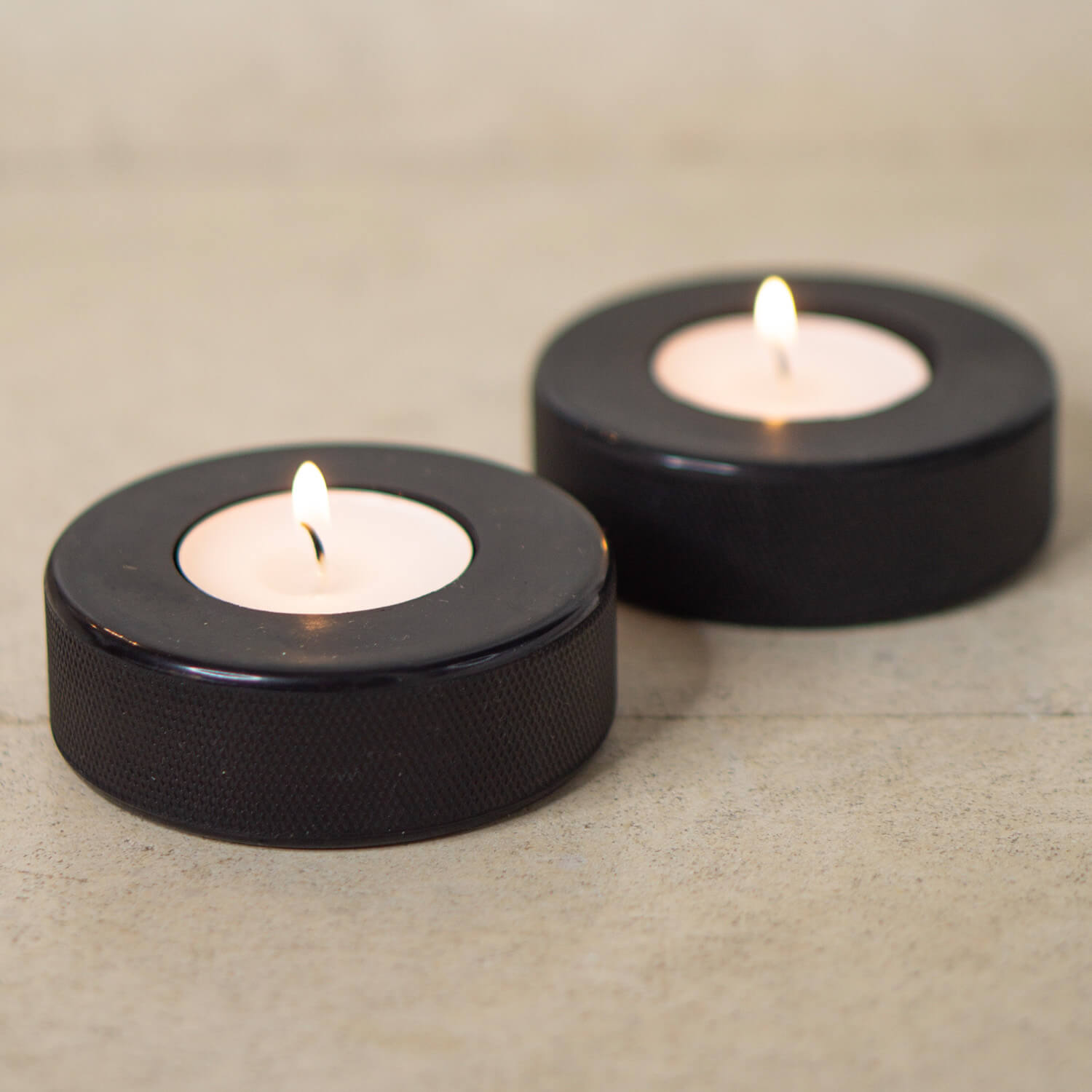 Zwei schwarze Teelichthalter aus Eishockey-Pucks mit brennenden Teelichtern auf hellem Untergrund