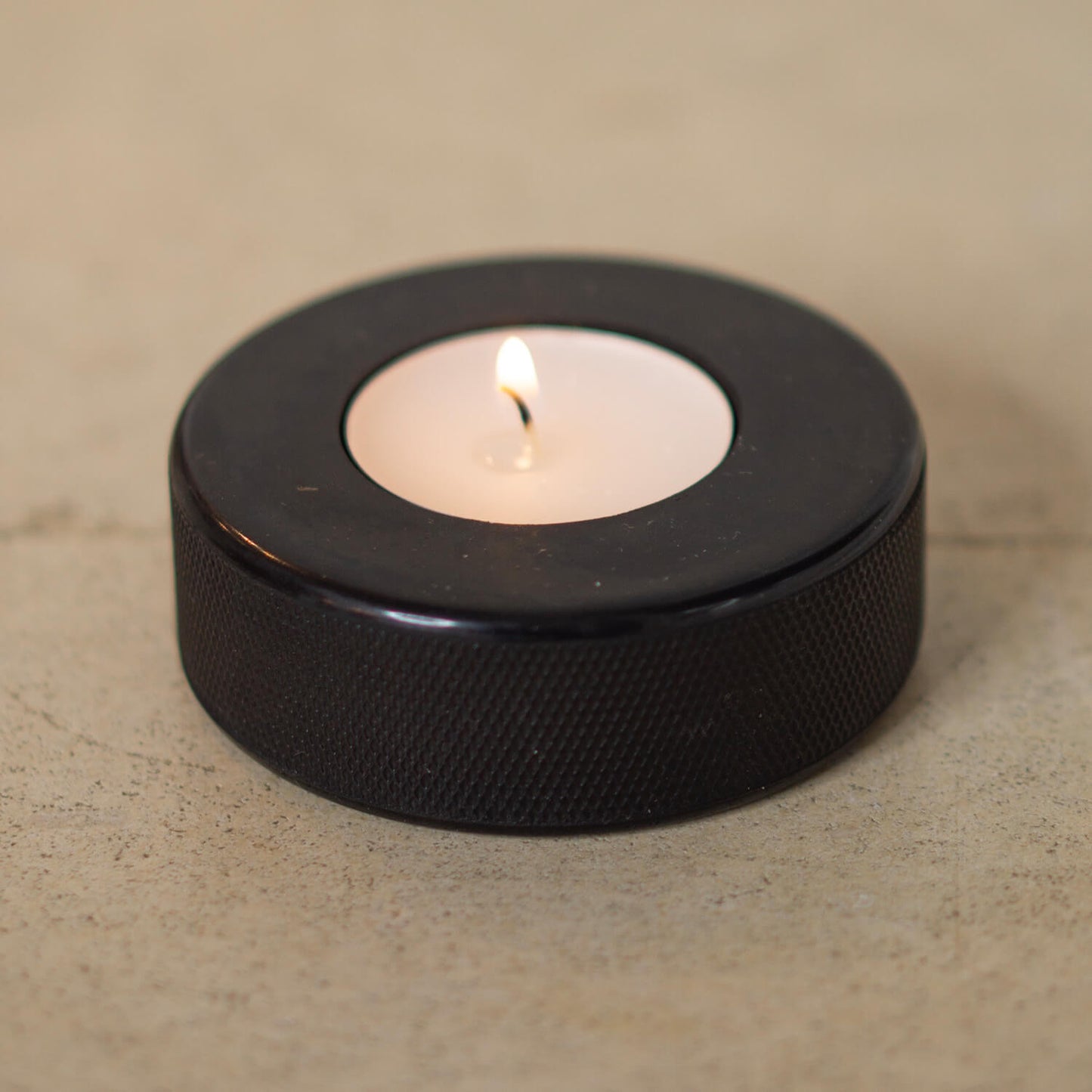 Nahaufnahme eines brennenden Teelichts, eingefasst in einen schwarzen Eishockey-Puck – minimalistisches Design trifft auf nachhaltiges Upcycling.