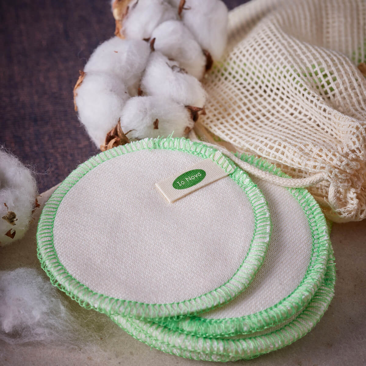 Wiederverwendbare Wattepads im 12er-Set von Io Nova – runde Baumwollpads mit grünem Rand, daneben ein Baumwollbeutel und frische Baumwollzweige auf hellem Untergrund.