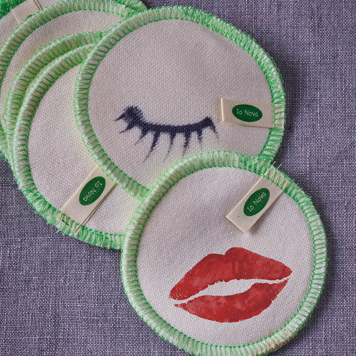 Wiederverwendbare Wattepads im 12er-Set von Io Nova – runde Pads aus Stoff mit grünem Rand, bedruckt mit Lippen- und Wimpernmotiven auf lilafarbenem Hintergrund.